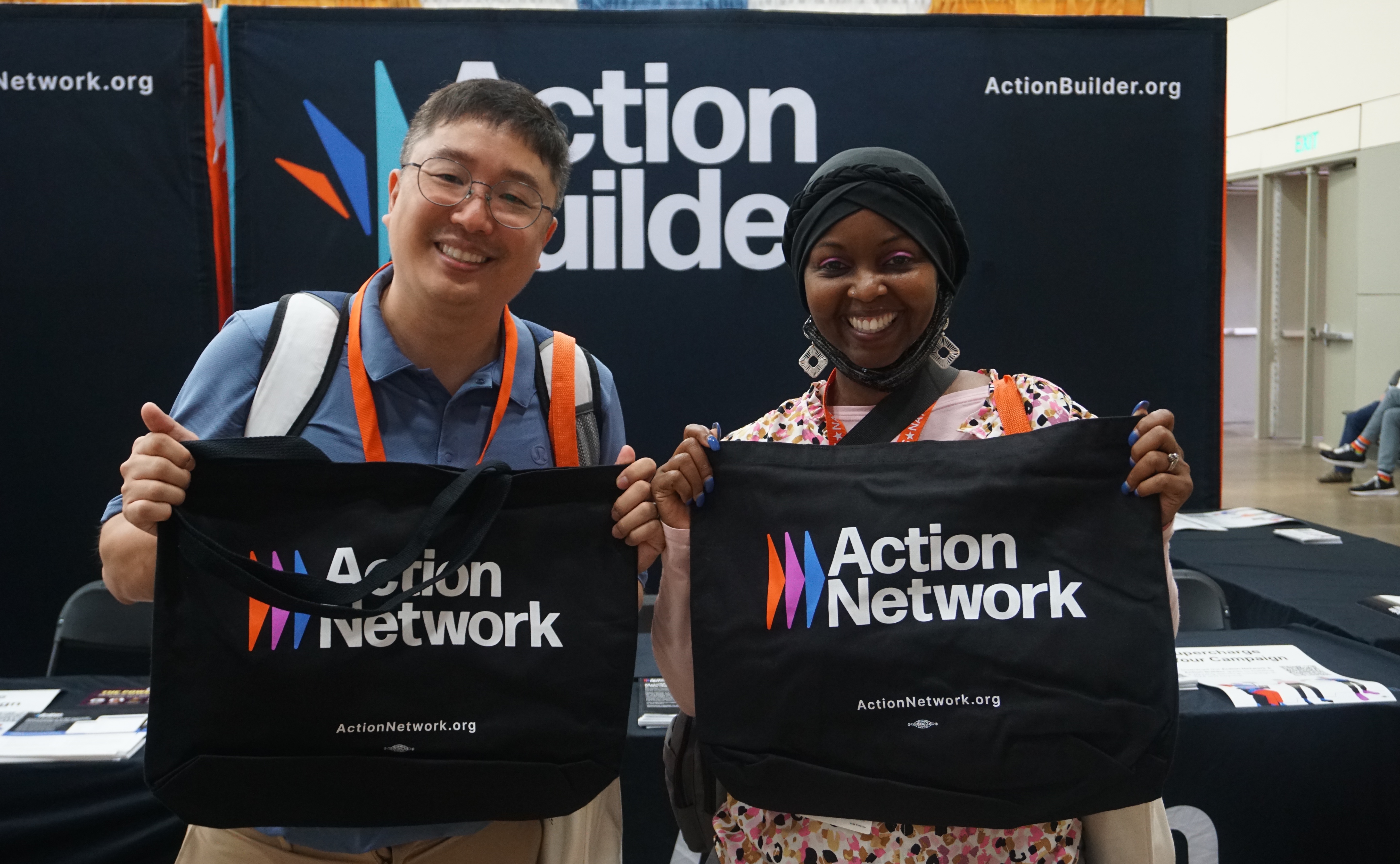Two team members from Asian Americans Advancing Justice-Atlanta standing in front of the Action Network booth holding up Action Network branded tote bags.