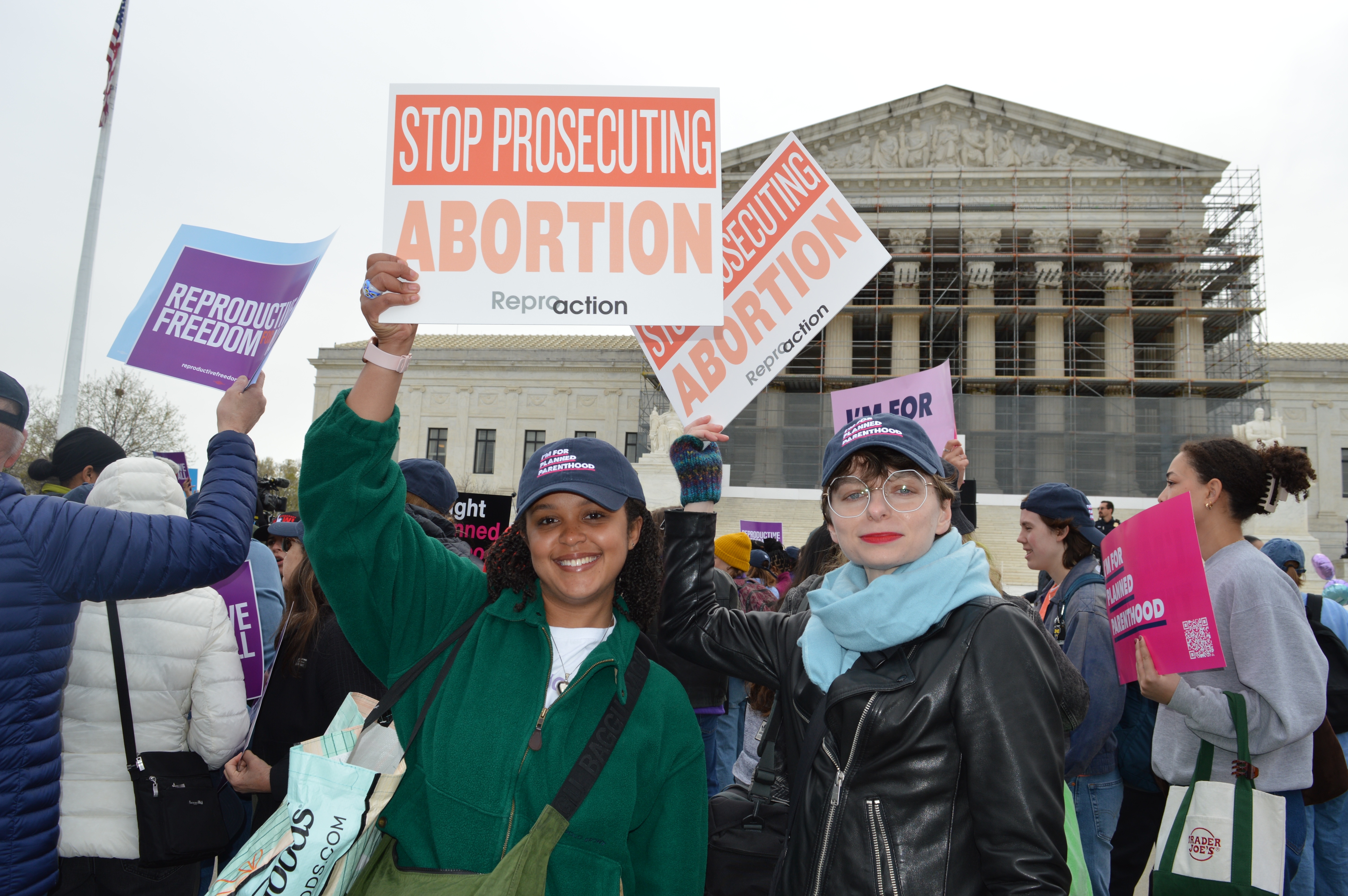 Two Reproaction employees stand in a crowd outside the Supreme Court smiling and holding signs that say Stop Prosecuting Abortion.