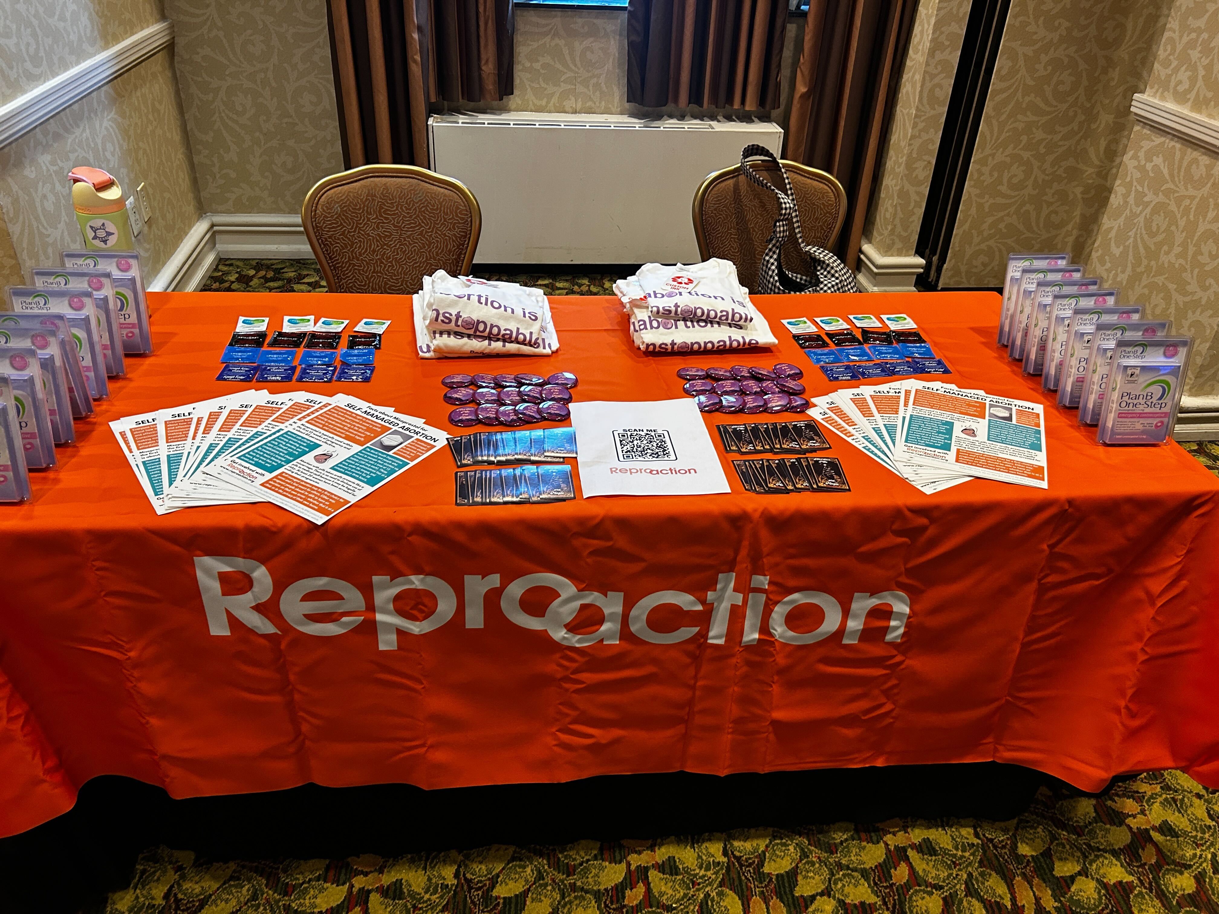 A table covered in an orange Reproaction table cloth, with flyers, stickers, shirts and other promotional materials.