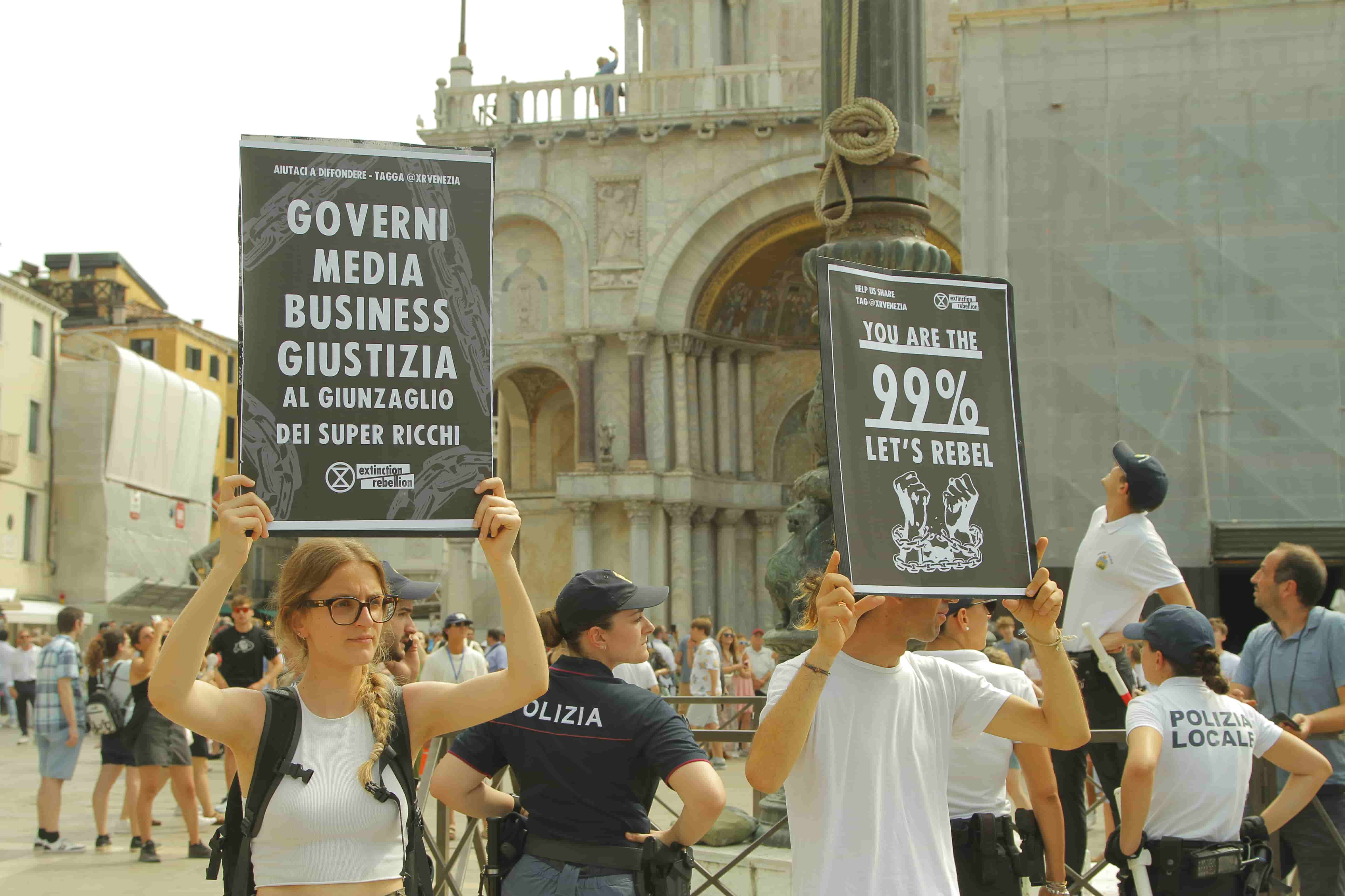 Photograph of a large but somewhat dispersed group of people in what appears to be a large city square surrounded by old-fashioned, ornate, stone buildings. The two people closest to the camera face toward the camera but without looking at it. They both hold black signs with white lettering--they look professionally printed. One sign says 