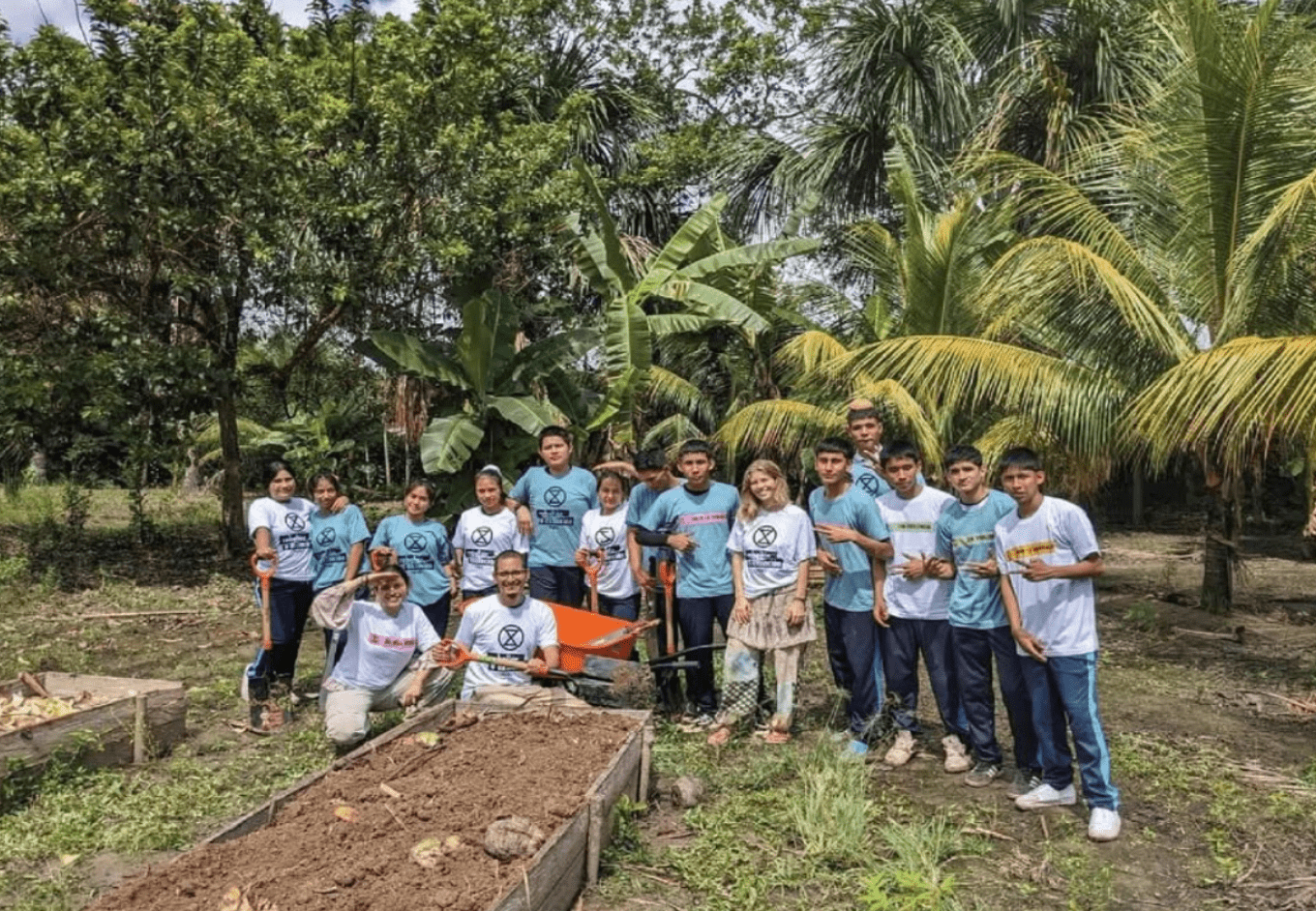 A photograph of a group of young people, perhaps older children or younger teens, clustered for a posted group photo behind some freshly-made garden beds. Do of the people are squatting in front of the others and may be adults or older teens, though it's hard to be sure. Some carry shovels. There is an orange thing among them that might be a wheel barrow, but the vie of it is partly blocked by the squatting people. Behind the people are palms, banana trees, and other trees and a climps of a partly-cloudy sky. Everything looks sunny and green and tropical. The people are all smiling, and most wear XR shirts. All are wearing short sleeves and long pants. One of them is wearing a skirt and pants.