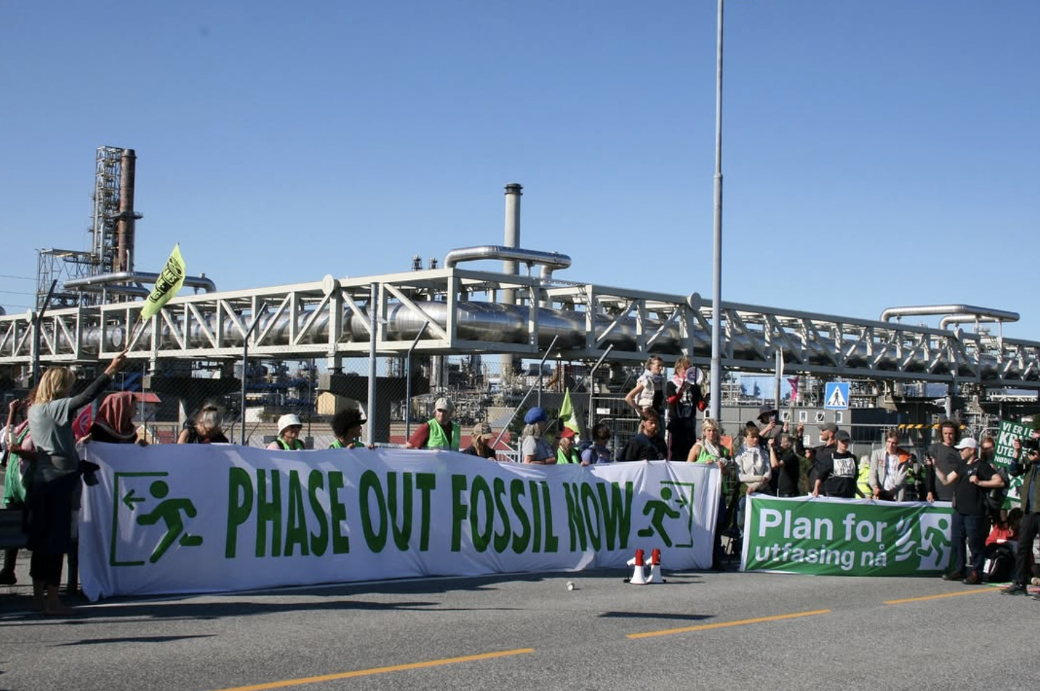 A photograph of a group of people standing in front of some kind of industrial complex dominated by both horizontal and vertical pipes. The people hold green and white banners, one of which reads 