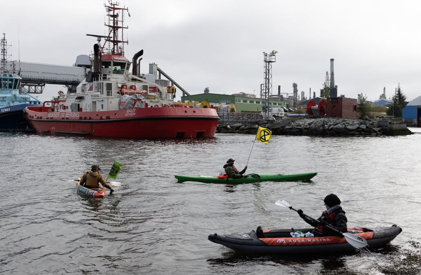 A photograph of what looks like a working harbor under a grey sky. The water is also grey. A large, red-hulled boat is visible in the background, as is part of a blue boat of similar size. In the foreground paddle three humans in kayaks, two of them holding small flags featuring the XR logo.