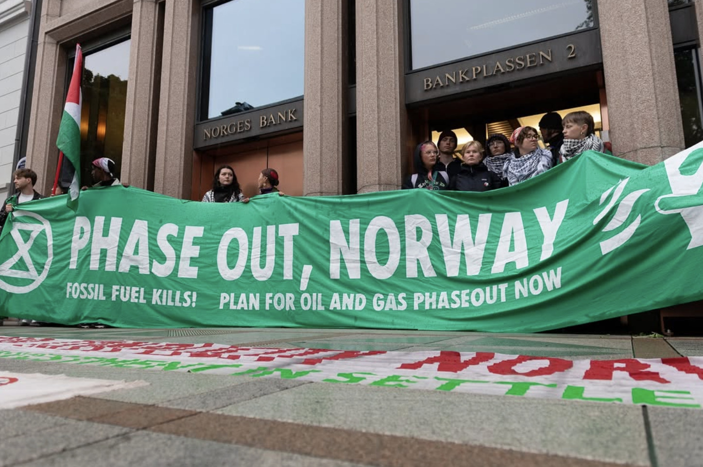 A photo of a small group of people holding a large banner in front of a building. Signs on the building read 