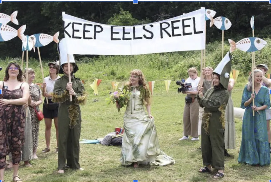 A photo of what looks vaguely like an outdoor wedding, though only a single bride is visible and no groom. The people standing on either side of the bride are wearing costumes that are probably meant to represent eels as they hold a banner above the bride that reads 