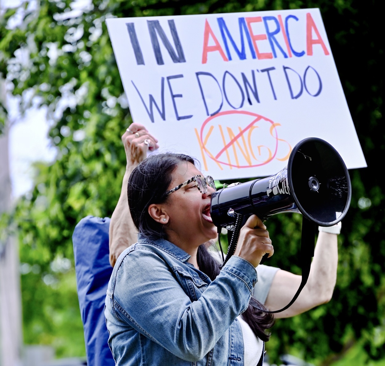 Rashida speaks into a bullhorn, standing in front of a sign that says In America We Don't Do Kings
