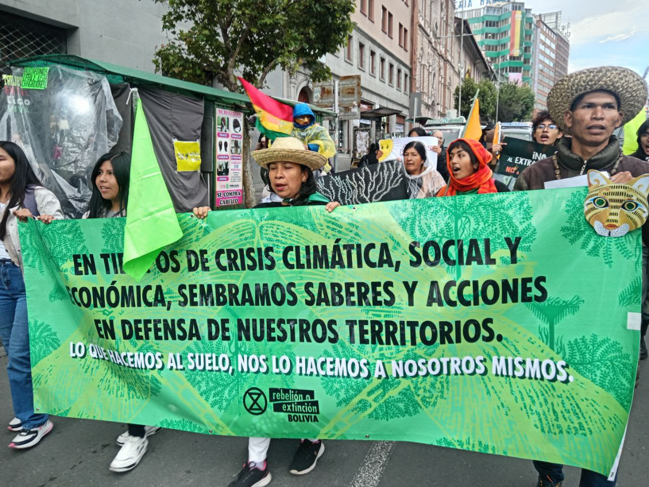 A photograph of a group of people in a protest march on a city street carrying various flags and signs. Those in front carry a large green banner with black and white lettering and the name and logo of Rebelion a Extinction Bolivia. Some of the words on the banner are obscured by fabric folds and another flag, making the message difficult to read, but it is in Spanish.