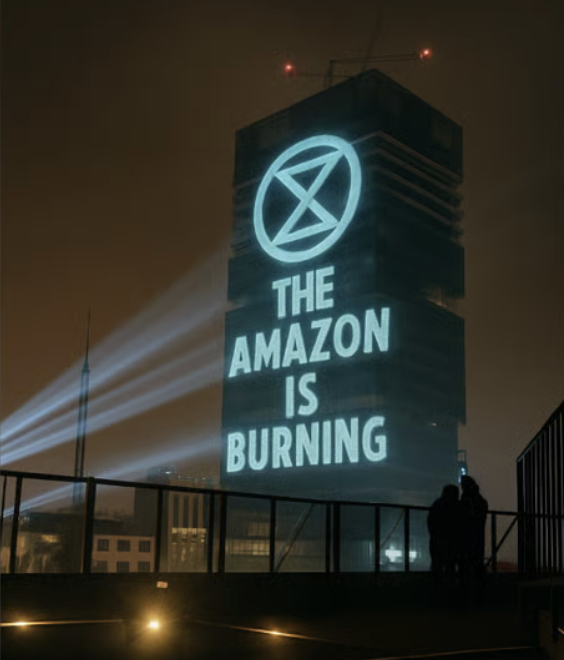 The image, which is in a realistic style but does not appear to be a photograph, shows a cityscape at night from what looks like the roof of a mid-sized building. The image is dominated by a building much taller than all the others on which is projected in greenish-white light the XR logo and the words THE AMAZON IS BURNING. On the roof in the foreground stand two people in silhouette apparently leaning together for comfort.