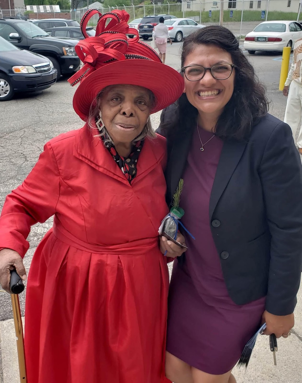 Rashida smiles with an older Black woman in a fantastic red hat, holding a cane.