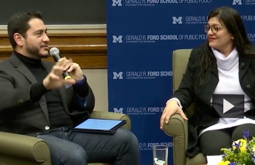 Abdul speaks into a microphone while seated at a panel. Rashida looks over at him, listening.