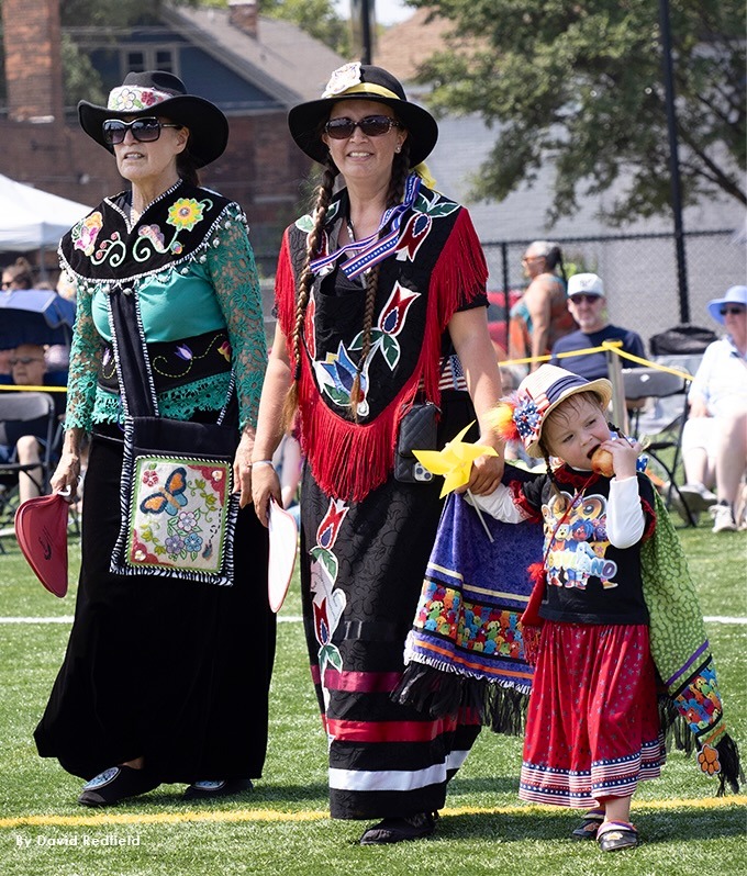 two native women and a child walk through a field in ceremonial clothes