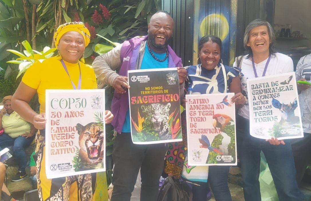 A photo of four people posing together, smiling and holding small posters with a lot of text. Other people are partially visible. There is vegetation in the background, but the setting appears to be indoors.