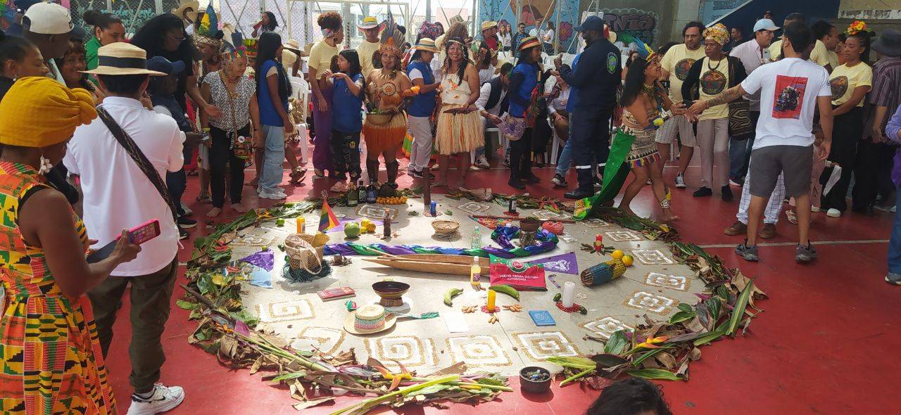 A photo of a group of people standing in a large room with a red floor. On the floor is a large, circular area perhaps three metres across defined by cut vegetation laid out almost like a big wreath, white inside with symbols and various objects laid out. It looks very ceremonial.