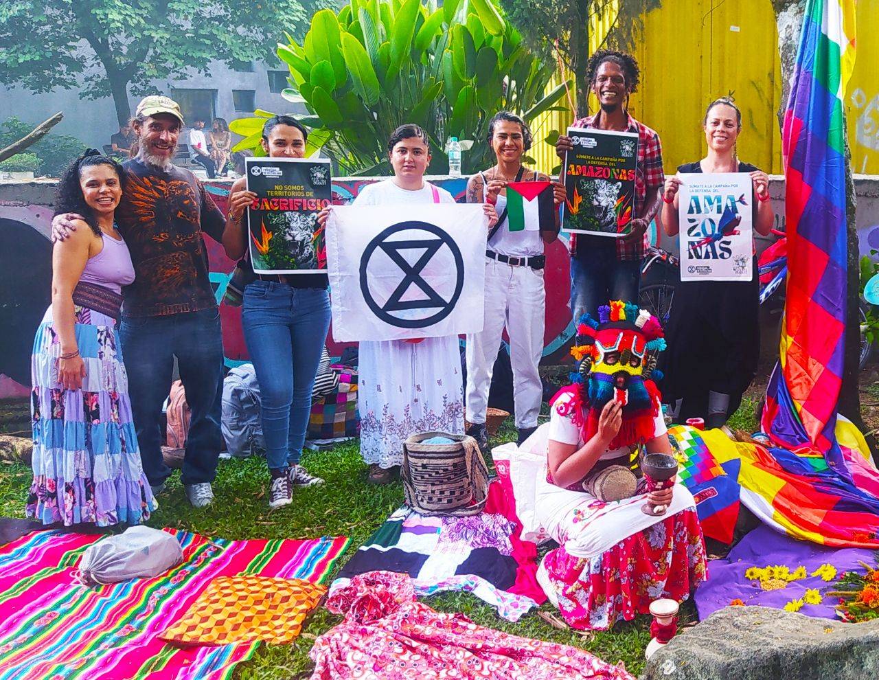 A photo of a group of people posing together outdoors. The vegetation is lush and green. There are buildings in the background, but the view of them isn't clear. Most of the people hold flags or signs, including an XR flag. There are brightly-colored blankets and the grass, and a large flag or quilt in bright colors hanging to the side. One person crouches in front of the other wearing a brightly-coloured mask and holding a cup.