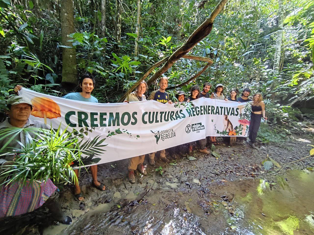 A photo of a group of people holding a long banner in front of a tropical forest edge. Water laps almost at their feet. The banner reads "creemos culturas regenerativas"
