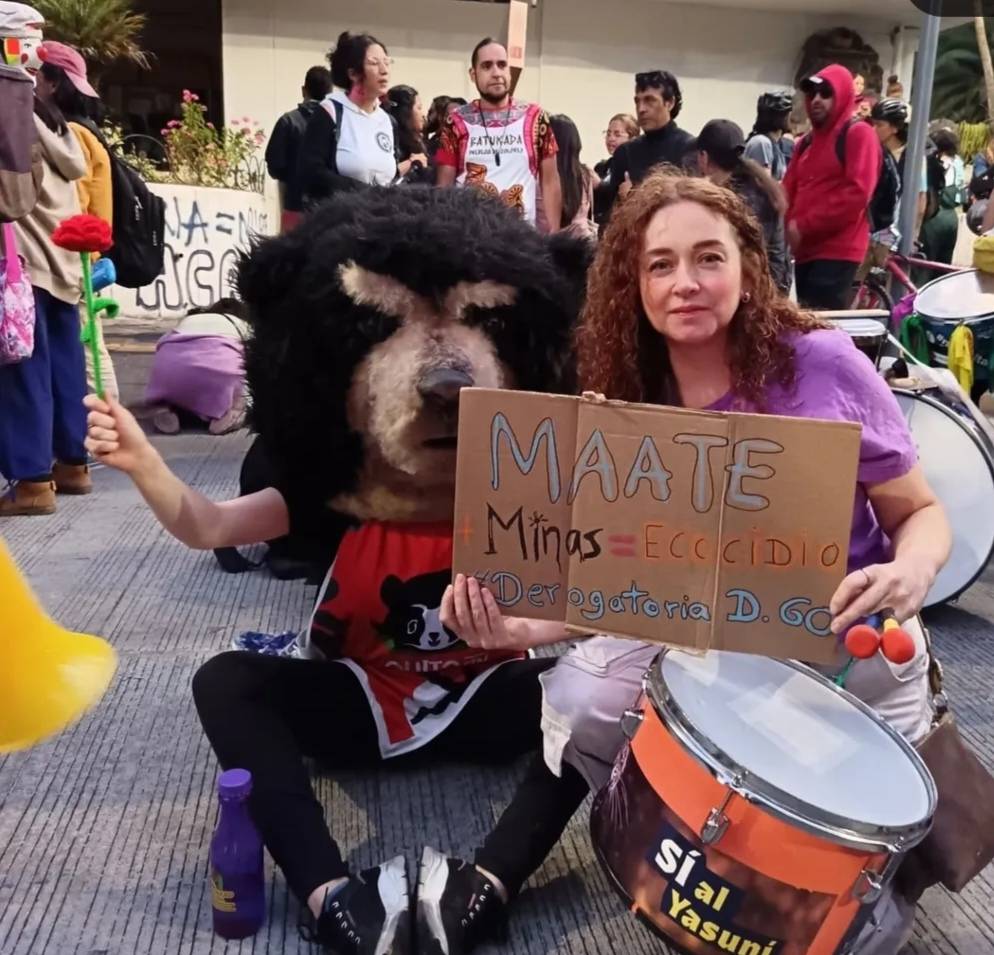 a photograph of a woman squatting and holding a cardboard sign and wearing a drum on a string around her waist. Beside her sits a person holding a fake red flower and wearing a very large bear head. They appear to be indoors. A crowd of people mill around behind them.