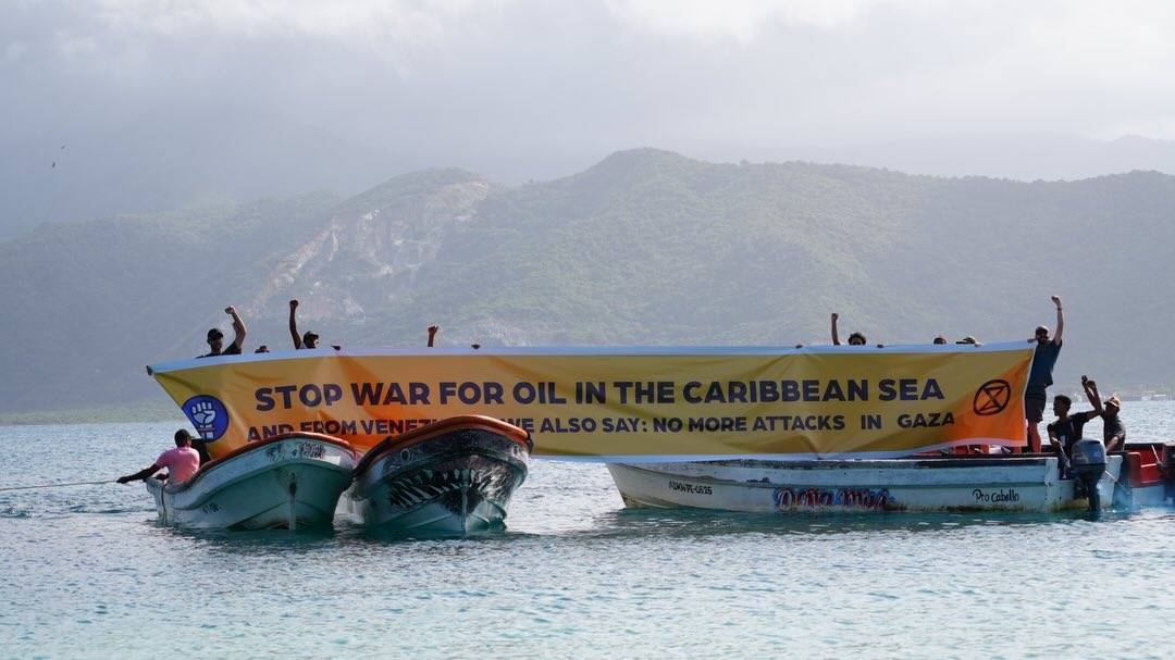 A photograph of three or four small boats in a tight group, the people on the boats holding up a yellow banner that reads "stop war for oil in the Caribbean Sea and from Venezuela we also say: no more attacks in Gaza". The banner has an XR logo and another logo of a white fist in a blue circle. Some of the people hold their fists up in the air. The seawater is relatively calm, the sky is cloudy, and in the background high, forested hills are visible.