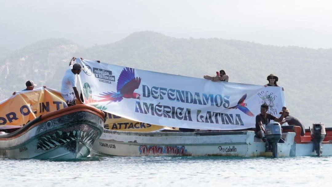 A photograph evidently of the same boats and people as the previous photo, but from a different angle and closer-up. The yellow banner is partially visible, though it seems to be being put away. Instead, the people are holding up a white banner that reads "defendamos America Latina" and features a color image of a scarlet macaw in flight and several logos.