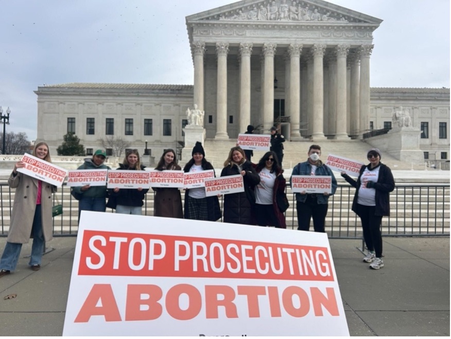 A group of people stand outside in DC. All holding signs that read Stop Prosecuting Abortion.