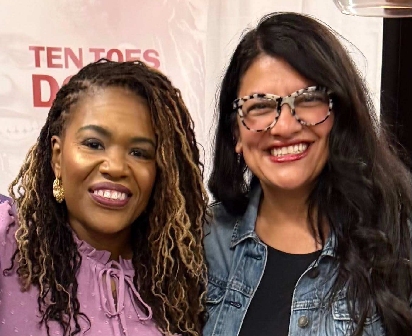 Cori Bush and Rashida Tlaib smile together for a photo at an indoor event, standing side by side.
