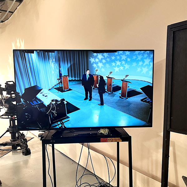 A photo showing a monitor backstage of the PBS Studios in which David Roth is visible standing with one of the producers as they prepare for the upcoming debate.