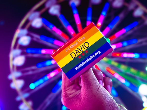 A photo of a campaign pin in the rainbow pride flag design that reads: “It’s about time. David. Roth for Idaho dot org.” The pin is held in someone’s hand up against a ferris wheel spinning at night.