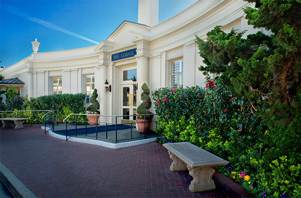 A photo of the front of The Lodge at Pebble Beach Resort. It is a white building with a curved front and marble columns and a lush garden in front.