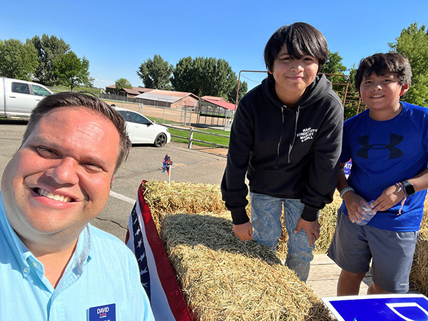 A photo of David Roth and his two sons. David is standing next to a pickup truck decorated for a campaign event with bales of hay in the back. David’s sons are standing in the bed of the pickup truck smiling at the camera.