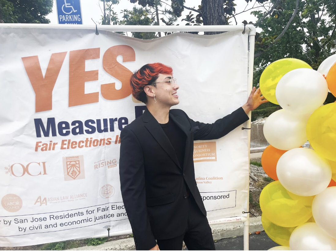 Image description: Neil is wearing a black suit and standing in front of a campaign banner that reads “Yes on Measure B.” He is looking into the distance with his arm extended. A white, yellow, and orange column of balloons is to his right.