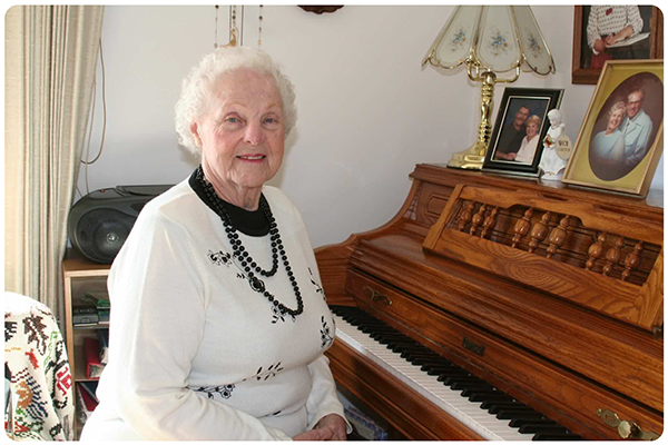Image of David’s Grandmother smiling contentedly while seated at her piano wearing a white long-sleeve shirt with black flowers and a black shirt underneath.