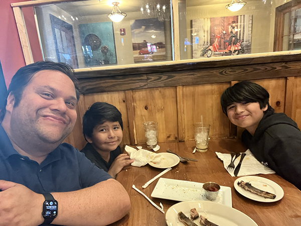 A photo of David Roth with his two sons seated at a restaurant table with empty plates in front of them, smiling contentedly as they just finished sharing a meal.