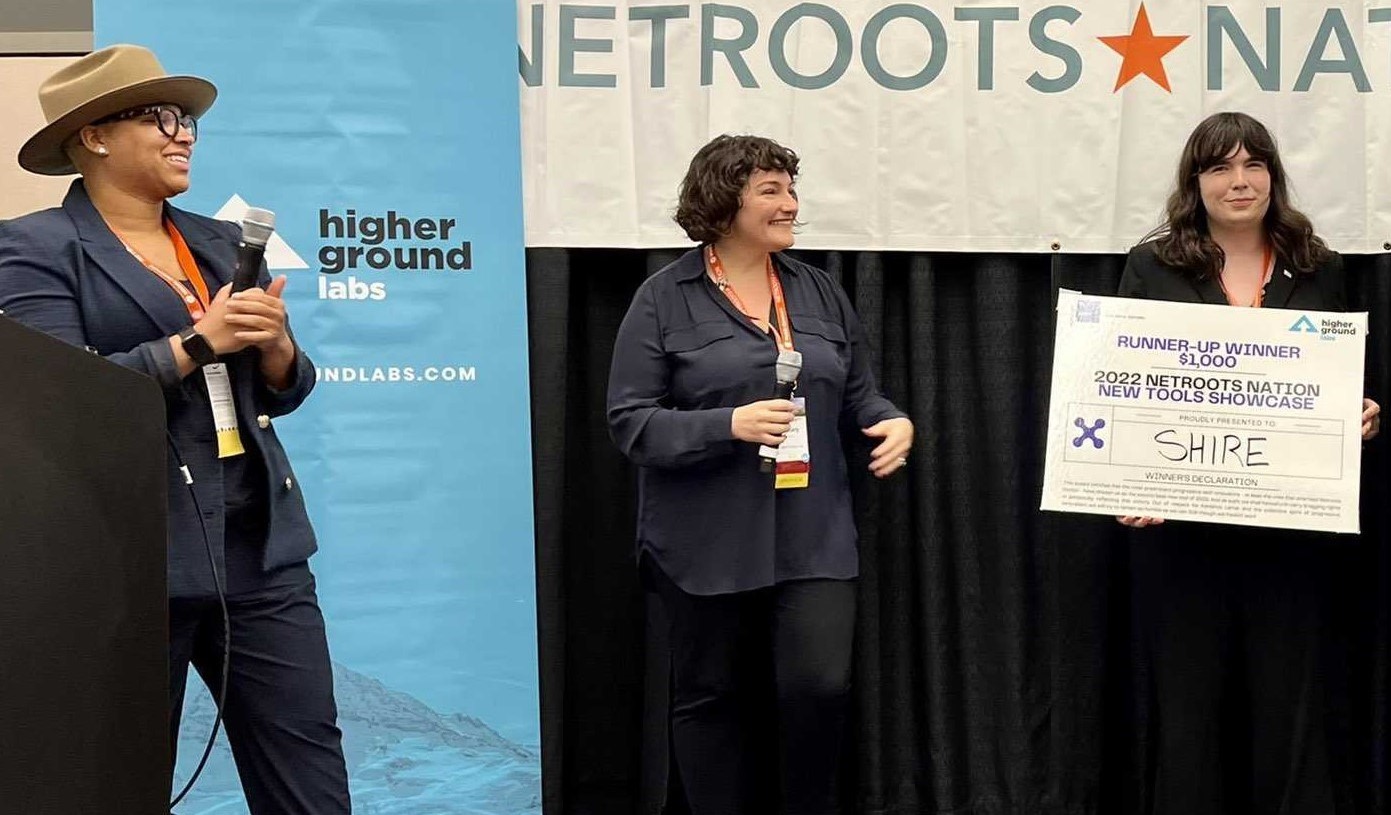 A photo taken at Netroots Nation depicting two women presenters awarding a prize to Shire. On the right, Shire founder Samantha Boucher is pictured holding a large check which says “Runner-up Winner $1,000, 2022 Netroots Nation New Tools Showcase, Shire”