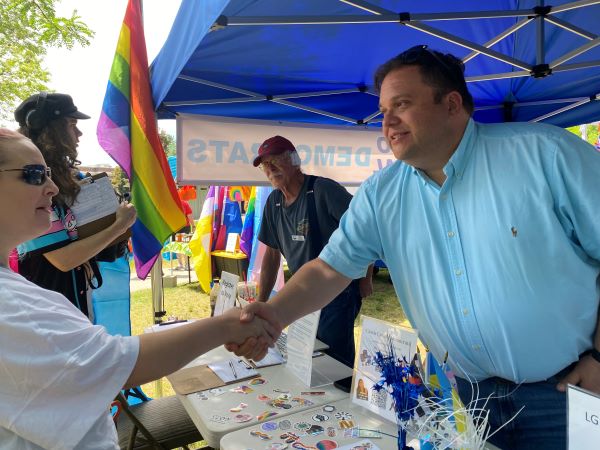 Photograph of candidate David Roth attending a Pride Festival. He is shaking hands and visiting with a festival attendant. There is a Pride flag waving in the background. Two other attendees visiting at the Roth campaign booth.