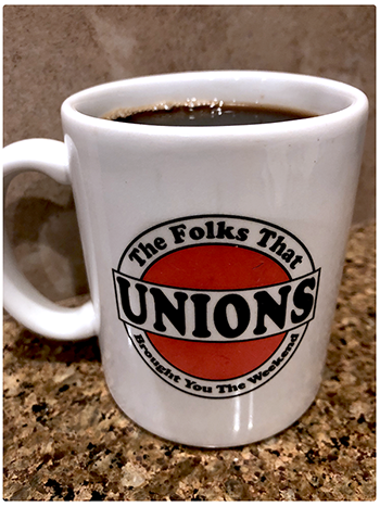 A photo of a white ceramic coffee mug with the words “Unions: The Folks That Brought You The Weekend.” The mug is sitting on a countertop and is full of coffee.