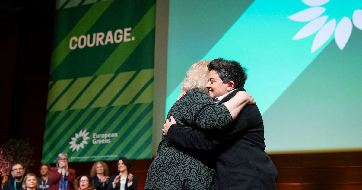 a photo of Mina Jack on stage at the European Greens Congress in Lyon hugging Jean Lambert. The word COURAGE on a banner behind them.