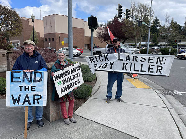 A photo of three protesters on a street corner in Washington’s 2nd congressional district. The man on the left holds a sign that says “End the War.” The woman in the middle holds a sign saying “Immigrants Built America.” And the man on the right holds a long banner that reads: “Rick Larsen: Baby Killer.”