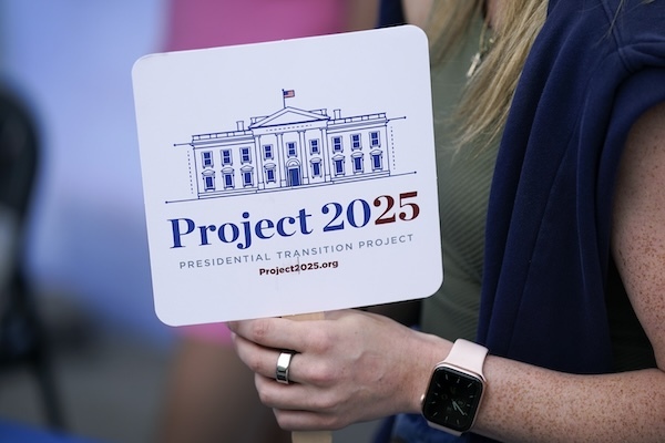 A close-up photo of a woman’s hand holding a small, white sign that reads “Project 2025 Presidential Transition Project.” Above the text is a digital drawing of the White House.