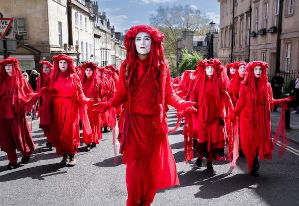 A photo showing people dressed in long bright red costumes with white-painted faces, with their hands outstretched and upturned