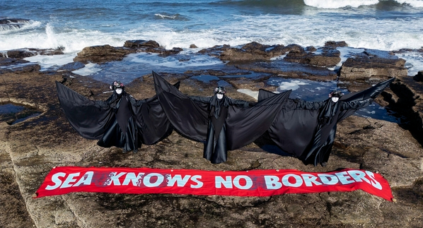 A photo showing three people dressing black robes standing on a rocky beach in front of a sign that reads Sea Knows No Borders