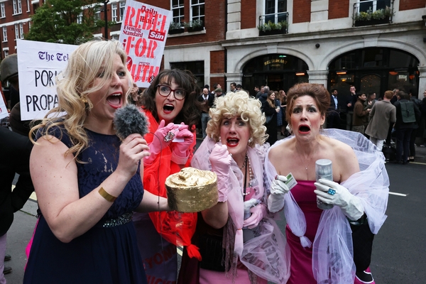 A photo showing four people dressed as women attending a glamorous party in the street. One is carrying a large gold fake pork pie and holding a microphone like a reporter