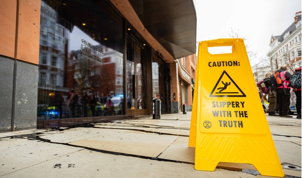 A photo of a street with the windows of an office on the left covered in black liquid, and in the foreground a yellow sign which is printed with Caution Slippery with the Truth