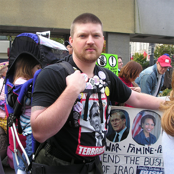 A photo of Jason wearing a t-shirt proclaiming George W. Bush an international terrorist, leaning on a protest sign identifying members of the Bush administration and their involvement in war crimes against the Iraqi people. Jason is wearing a backpack-style carrier with his daughter as a toddler seated inside.