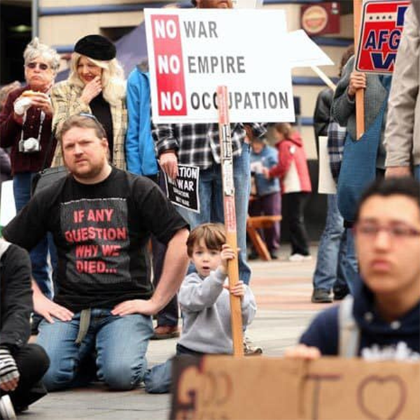 A photo of Jason wearing an anti-war t-shirt and kneeling on the ground at an anti-war protest. His son as a young boy is kneeling beside him holding up a sign that says “No War, No Empire, No Occupation.”