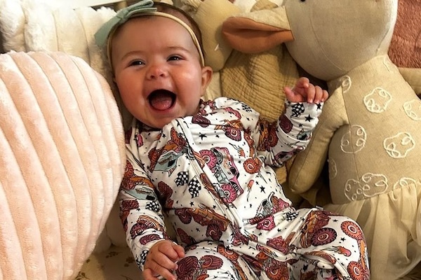 A photo of a beaming 4-month-old Tanna wearing a tractor onesie and a blue bow on her head.