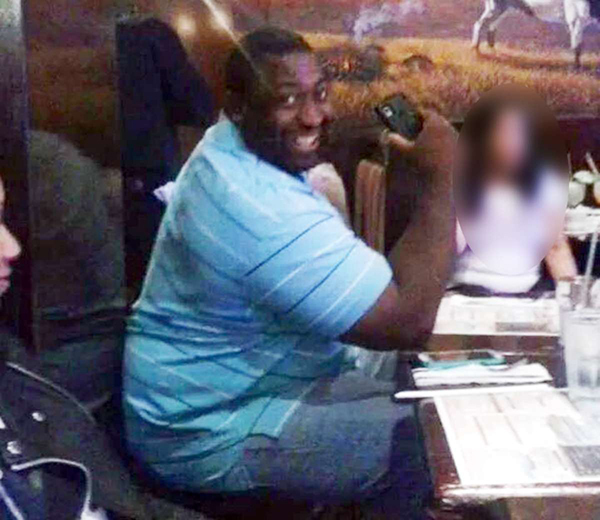 A photo of Eric Garner at a restaurant with family. He is wearing a blue striped polo shirt and jeans, holding his cell phone as he leans on this elbows on the table, smiling exuberantly at the person taking the photo.