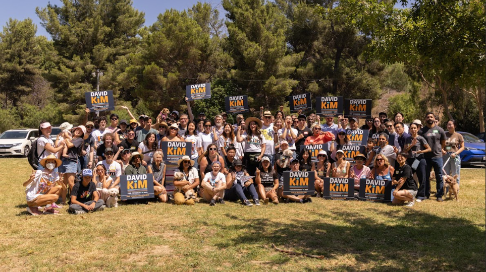 A photo of the crowd of supporters gathered for David Kim’s community picnic with a large group holding campaign signs on a bright sunny afternoon in the park.