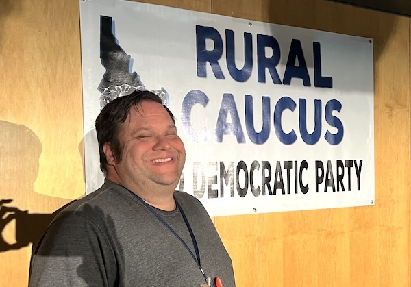 A photo of David grinning ear to ear standing in front of a sign that reads “Rural Caucus, Idaho Democratic Party.”