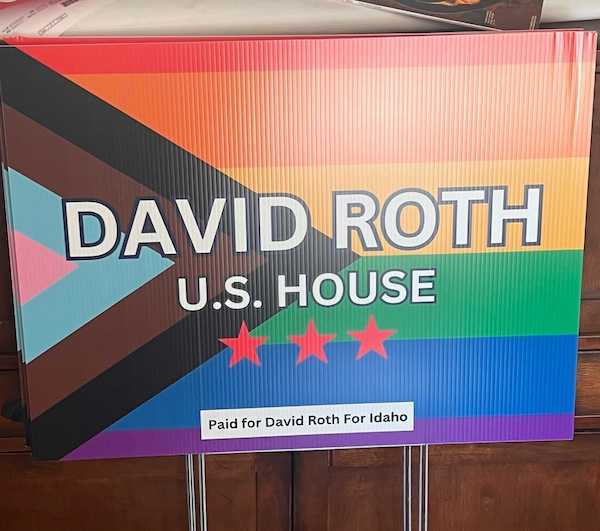 David Roth U.S. House campaign yard sign featuring the Progress Pride flag in the background. The sign reads 'David Roth U.S. House' with three red stars underneath. At the bottom, a small white rectangle contains the text 'Paid for David Roth For Idaho.' The Progress Pride flag colors include rainbow stripes with a chevron design on the left in brown, black, white, pink, and blue.