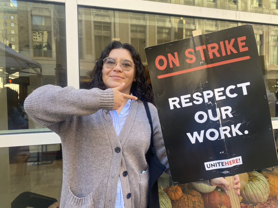 A photo of a striking worker from the UNITE HERE strike line holding a sign that reads, “On strike. Respect our work,” above the UNITE HERE logo.