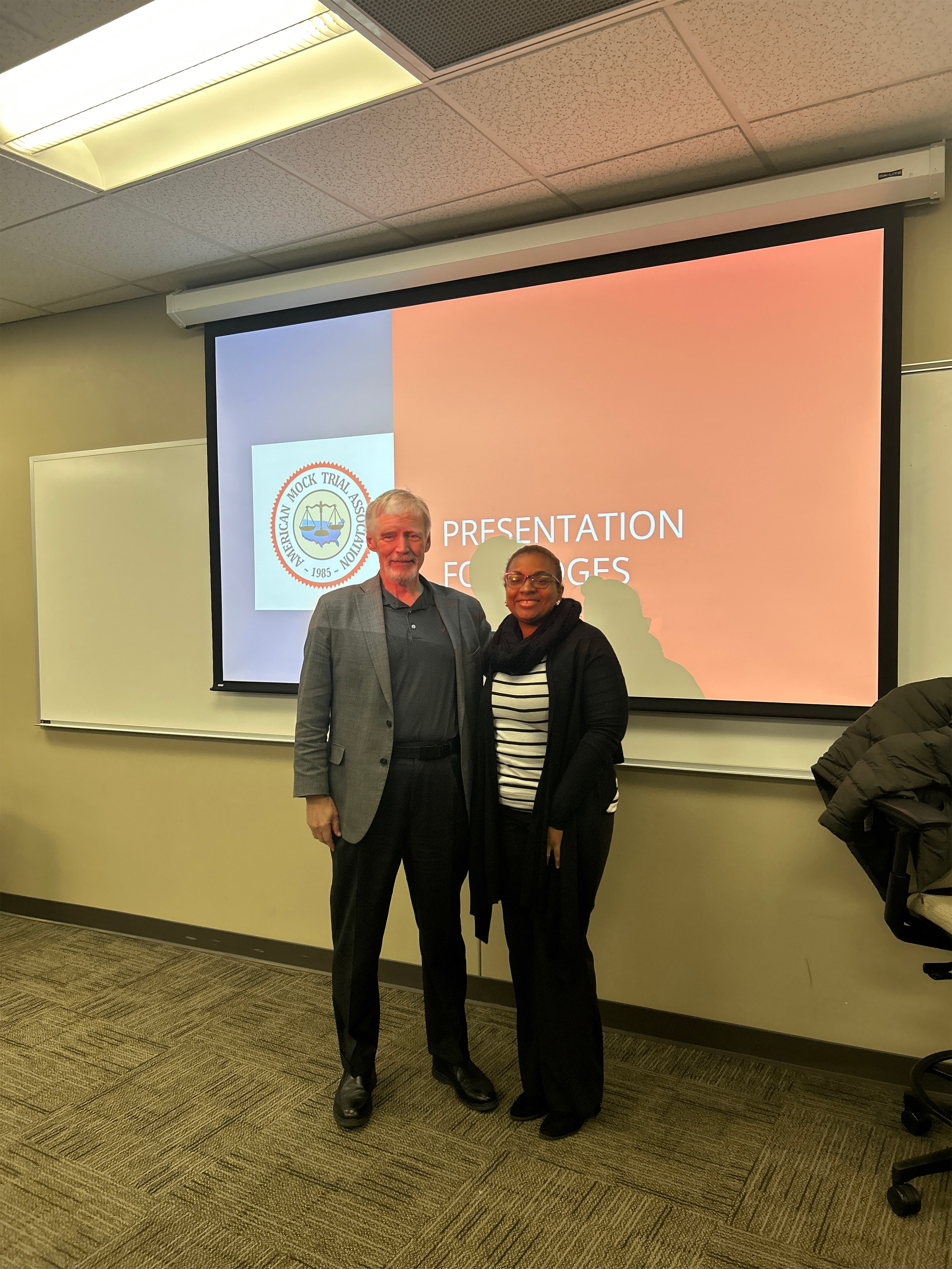 A smiling Larry and Santita stand in front of a projector screen at the Crimson Classic Mock Trial Competition.